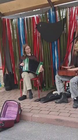 The video shows two young people performing music outdoors under a covered structure. The person on the left, wearing glasses and a striped sweater, plays an accordion while seated on a red chair. The person on the right, with green-dyed hair and wearing a black shirt and grey pants, plays a stringed instrument that appears to be a ukulele or a small guitar, also seated on a red chair. A large black cut-out of a bird is mounted on a backdrop of colorful hanging ribbons behind them. In the foreground, to the left, is an open red and purple instrument case. The ground is paved with brick-like stones, and foliage is visible to the left. The overall scene suggests a casual outdoor performance.
