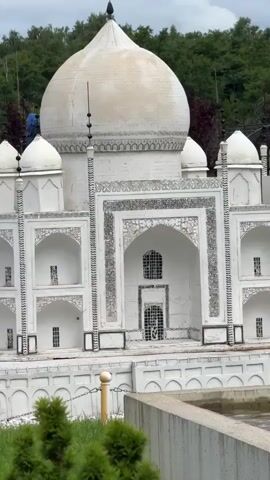 A miniature replica of the Taj Mahal is shown in an outdoor setting. The structure is made of white material and features a large dome, four minarets, and intricate architectural details. It is surrounded by a garden with green grass, manicured bushes, and trees in the background. A water feature with fountains is visible in front of the replica. The sky is cloudy, with patches of blue peeking through. There are no people clearly visible interacting with the replica, though a blue object, possibly a piece of clothing or equipment, is perched on top of one of the minarets on the left side of the structure. The camera slowly pans out, revealing more of the surrounding park-like environment.