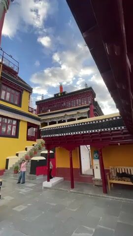 The video pans across a courtyard of a building complex that appears to be a monastery or temple. The architecture is vibrant, with brightly painted walls in yellow and red, accented with intricate decorative elements. A tall flagpole with prayer flags stands prominently in the center of the courtyard. Several people are visible, some walking, some standing, and one person is seated on a low wall. The sky above is a bright blue with scattered white clouds, suggesting pleasant weather. The ground is paved with stone tiles. The overall impression is of a cultural or tourist site with active visitors.
