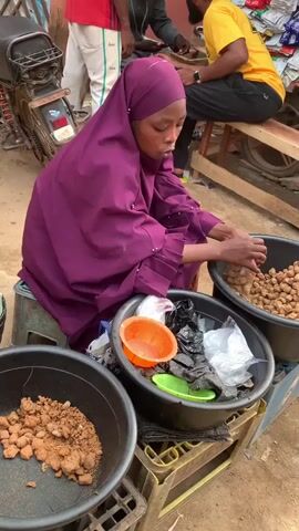 A young woman wearing a purple hijab and matching robe sits on a blue and yellow plastic crate outside. In front of her are three black plastic tubs. The tub to her left contains a pile of rough, brown lumps. The tub in the center contains an orange plastic bowl, a green plastic plate, black pieces of charcoal, and clear and black plastic bags. The tub to her right contains a pile of round, tan pieces. The woman reaches into the tub in the center, pulling apart a black plastic bag. In the background, other people are standing or sitting on a wooden bench near a motorcycle and a vendor's stall.