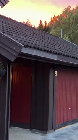 The video shows a close-up, static shot of a dark-tiled roof of a building. The building appears to be a garage with two red doors, one on the left and one on the right. A house number "2" is visible on the right door. Above the roof, a dramatic sunset unfolds with vibrant hues of pink, orange, and red filling the sky. Silhouetted trees and what appear to be evergreen trees are visible against the colorful sky. The lighting suggests it is dusk.
