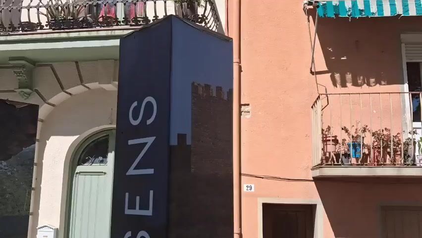 The video shows an outdoor scene, likely in a European town or village. The camera focuses on a black rectangular pillar with the word "QUESENS" printed vertically in large, white letters. The pillar appears to be part of a tourist information system, with smaller text and images related to local attractions. To the left of the pillar is a white building with a light green door, and to the right is a peach-colored building with a balcony decorated with potted plants. The street is paved with gray tiles. The number "29" is visible on the peach building. There are other similar pillars in the background, with the words "Santa Julia dels Corts" visible on one of them. The scene is bright and sunny.