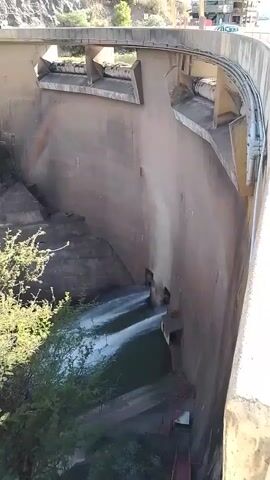 The video shows a view from above of a concrete dam. Two streams of water are flowing through openings in the dam wall into a pool below.  A series of steps lead down the wall towards the water.  The top of the dam forms a bridge, and pipes run along the side of the dam.  Green plants grow at the base of the wall.  Buildings and a hill are visible in the background.  The lighting suggests it is daytime.