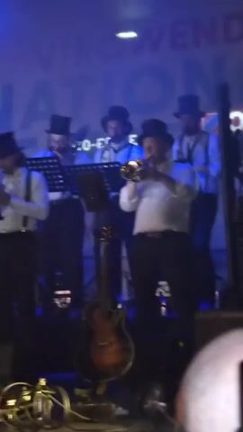 A band plays on a stage under blue and white lights. Five members of the band are wearing black top hats, white shirts, and suspenders. One band member in the center plays a brass instrument, possibly a trumpet. To the left of the stage, another person in a sparkly jacket and yellow pants plays a guitar. In the foreground, the heads of audience members are visible, with one person holding up a phone. The background has a sign with blurry white text. The video also briefly shows a street scene at night with people holding small fire torches.