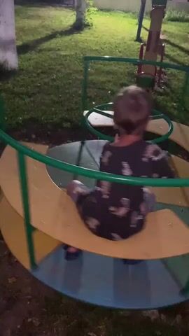 A young boy with short brown hair is sitting on a merry-go-round at a playground. He is wearing a dark t-shirt with a tie-dye pattern and grey shorts. The merry-go-round has yellow and green seats and a green metal frame. The boy is holding onto the steering wheel and appears to be spinning himself. The ground around the merry-go-round is covered in grass and dirt. In the background, there is a swing set and some trees. The lighting suggests it is evening.