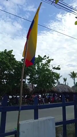 The video opens with a Colombian flag flying from a wooden pole. The flag is positioned on the left side of the frame. Behind the flag, there are several thatched-roof structures, possibly restaurants or beach huts, and lush green trees. A blue fence runs across the middle of the frame, separating the foreground from the background. In the foreground, there is a white concrete structure and part of a wheelchair is visible on the right. The camera then pans to the right, revealing a white brick building with signage that reads "Hotel-Rest. WAIKIKI". The building has a red and yellow circular logo to the left of the text. Power lines and a utility pole are visible above the building and the thatched-roof structures. The sky is cloudy, with patches of blue. People are visible sitting at tables in the background, under the thatched roofs. There are also a few motorcycles parked in the background. The video appears to be filmed outdoors, likely in a tropical or coastal setting.