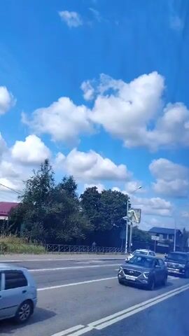 The video shows a view from inside a moving vehicle, looking out at a highway on a clear day with white clouds in a blue sky. There are several cars and trucks on the highway, traveling in both directions. Some of the cars are passenger vehicles, while others are larger trucks, including a red dump truck and a white semi-truck. There are also trees and buildings visible along the side of the road, as well as a billboard. The video appears to be filmed from a side window of the vehicle.