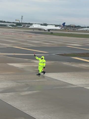 A ground crew member, clad in a bright lime green high-visibility suit with the hood up, is captured actively marshalling an aircraft on a wet airport tarmac. The individual holds two red/orange signal wands, one raised high in a signaling gesture and the other lowered. They wear black boots, appropriate for the operational environment.

In the background, a white United Airlines jet, identifiable by the "UNITED" text on its fuselage and the blue tail with the globe logo, is positioned on a taxiway. The aircraft appears to be a twin-engine passenger plane, likely an Embraer E-Jet.

The concrete surfaces of the apron and taxiways are visibly damp or wet, indicating recent rainfall or overcast, moist conditions. The sky above is uniformly gray and overcast. In the further background, other smaller aircraft, airport buildings, and utility structures like white storage tanks are discernible, confirming an active airport setting. The scene takes place during daylight hours. The overall atmosphere suggests a typical operational day at an airport, possibly in San Antonio, United States, under less-than-ideal weather conditions.