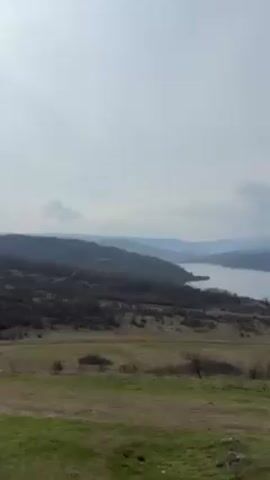 The video pans across a wide landscape, starting with a view of a large body of water, presumably a lake or reservoir, surrounded by rolling hills and mountains in the distance under a cloudy sky. The foreground features a grassy field with sparse vegetation. As the camera moves, it reveals a small group of people walking towards a rocky outcrop. A single individual then sits alone on the rocks, overlooking the expansive view. The camera continues to pan, showcasing the rocky terrain and the lake below before finally pointing up at the cloudy sky.