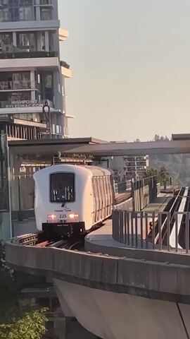 A white automated train labeled 225 with "TRANSIT LINK" branding travels on an elevated track. The train has its headlights on. A modern building with multiple windows is visible on the left side of the frame, with text on it that partially reads "HIGHPQINT RYLERMAC.COM". The sky is clear and bright, suggesting daylight. The train appears to be moving away from the camera, curving to the right along the track. The track is bordered by a grey concrete barrier on the left and a metal fence on the right. Trees are visible in the background. The scene appears to be an urban environment with modern architecture.