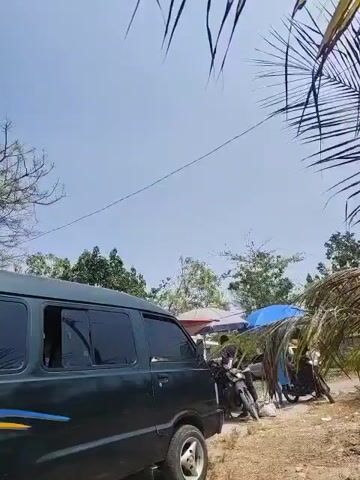 A dark green van is parked on a dirt road. The van has tinted windows and a yellow and blue stripe on the side. In the background, there are trees and two motorcycles parked under umbrellas. The sky is clear and blue.