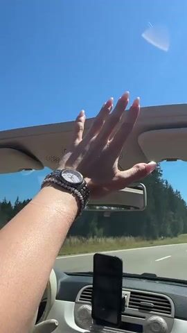 A first-person perspective video shows a woman's hand, adorned with bracelets and a watch, reaching up towards the roof of a convertible car. The hand is positioned in the upper right quadrant of the frame. The car's interior is visible, including the dashboard with a phone mounted on a holder. The background shows a clear blue sky and a tree-lined road indicating movement. The woman's fingernails are painted pink.