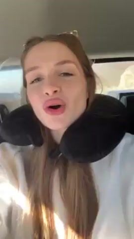 A young woman with light brown hair and wearing a white sweatshirt and a black neck pillow is in a car. She is facing the camera, and the car appears to be moving, with scenery visible outside the windows. The woman is singing along to music, with her mouth open wide. The light is bright, possibly from the sun shining through the windows.