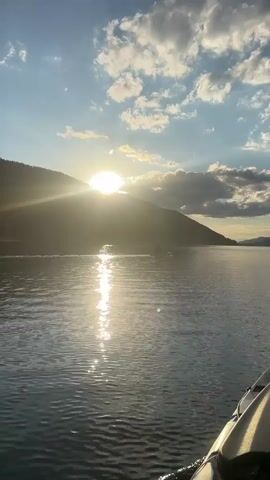 The video captures a serene view of a lake at sunset from the perspective of a kayak. The sun, partially obscured by a mountain range, casts a brilliant golden glow across the water, creating shimmering reflections. The sky is a blend of blue and white, with scattered clouds catching the warm light. In the distance, a small boat or jet ski can be seen moving across the lake. The foreground shows the front portion of the kayak, hinting at a peaceful journey on the water.