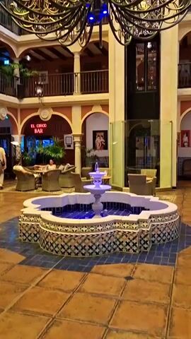 The video shows the interior of a building with a central atrium. A large, ornate chandelier hangs from the ceiling, which is made of yellow-tinted glass panels. In the center of the atrium, there is a decorative fountain with blue lighting. Around the fountain, there are seating areas with upholstered furniture. In the background, there is a bar with a sign that reads "EL CIRCO BAR." There are balconies with railings on multiple levels overlooking the atrium. Several people are visible sitting in the seating areas and walking around. The architecture appears to be inspired by Spanish or Mediterranean styles, with arched doorways and tiled surfaces.