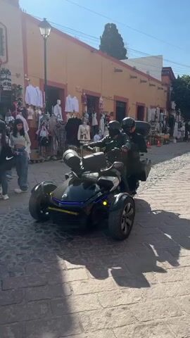 The video shows a stationary black Can-Am Ryker three-wheeled motorcycle, with two people on it, parked on a cobblestone street. Both riders are wearing helmets and dark clothing. The driver appears to be wearing a leather jacket and a dark face covering. Shops line the street behind the vehicle, displaying clothing and other goods. Several pedestrians are walking nearby, including a young woman in jeans and a crop top. The buildings are painted in warm colors, and the day appears sunny.