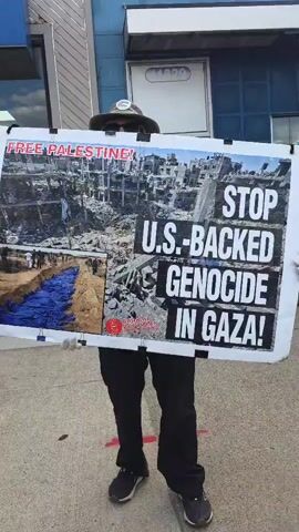 A person stands on a sidewalk holding a large sign that says "FREE PALESTINE! STOP U.S.-BACKED GENOCIDE IN GAZA!" The sign also includes images of what appears to be bombed-out buildings and a group of people standing near what appears to be a mass grave. The person is wearing a hat, sunglasses, black pants, and dark sneakers.
The camera moves to show a small group of people standing near the entrance of a store with signs that say "We Accept EBT & WIC" and "Halal Meat Available". A man is sitting on the ground in front of the store. The camera pans out to show a larger group of people protesting. Some people are holding signs, and others are wearing masks. The building has a blue awning and a sign that says "ROYAL" on it. The sky is blue with some clouds. The protest appears to be taking place in front of the store on a city sidewalk.