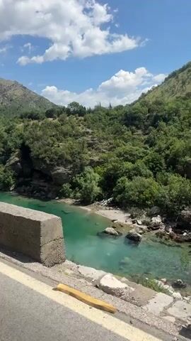 The video shows a scenic view of a river and mountains. The river is a clear turquoise color and flows between lush green hills. The sky is blue with fluffy white clouds. The video is taken from what seems to be a roadside. The wind noise is audible, and the camera pans upwards slowly. There are no people visible in the video.