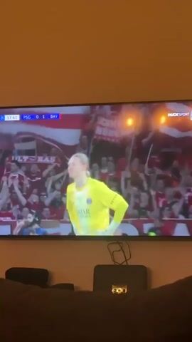 A flat-screen television displays a live soccer match in what appears to be a living room setting, with a brown couch visible in the foreground. On the TV screen, a male soccer player, likely a goalkeeper, with long blonde hair tied back, is shown wearing a bright yellow jersey. The jersey features the Paris Saint-Germain (PSG) logo and "QATAR AIRWAYS" as a sponsor. The player has a focused and intense expression. In the background on the TV, a crowd of fans, predominantly in red and white, can be seen in a stadium setting.

The on-screen scoreboard indicates the match is between PSG and BAY (Bayern Munich), with Bayern leading 0-1. The game time displayed is 17:42. The channel or broadcast information "M4XSPORT 3 LIVE" is visible in the top right corner of the screen. Below the television, a black electronic device with a few small lights and some cables is placed on a surface. The video was captured in Essen, Germany.