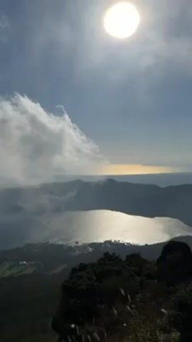 The video presents a scenic view from a high vantage point, possibly a mountaintop. The sun shines brightly in the sky, partially obscured by thin clouds. Below, a lake reflects the sunlight, creating a shimmering effect on its surface. Mountains surround the lake, some partially hidden by clouds. Vegetation, including dark rocks and tall grass, is visible in the foreground.
