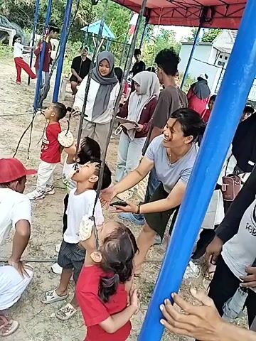 The video shows a group of people, mostly children and young adults, participating in a cracker-eating contest. The children are standing in a line, each with a cracker suspended from a string in front of their mouths. They are attempting to eat the cracker without using their hands. An adult male in a red cap is holding the string for one of the children, while other adults watch and encourage the participants. The event appears to be outdoors, with a tent set up in the background and trees visible. The ground is covered in dirt and sand.
