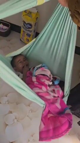 A baby is lying in a light green fabric hammock. The baby is wrapped in a pink and white blanket with a pattern. The baby is looking up and appears to be crying or about to cry. There is a patterned wall behind the hammock and a yellow box on a shelf in the background. The hammock is suspended from above.