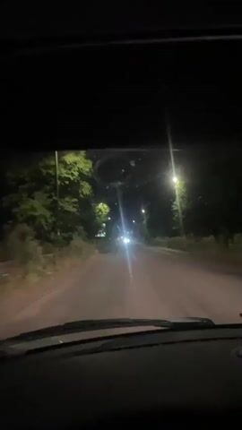 The video appears to be recorded from inside a car at night. The road ahead is lit by streetlights and the headlights of oncoming vehicles. Trees line the sides of the road. Inside the car, the dashboard lights are visible, and a phone is mounted on the windshield. A person's hand is on the steering wheel. In the later frames, other passengers are visible in the car, and the camera is moving erratically, suggesting the car may be in motion. The lighting is dim, and the focus is inconsistent.