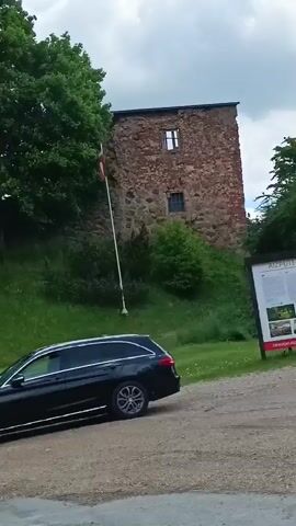 A black Mercedes-Benz C-Class station wagon drives from right to left on a dirt road. In the background, a green grassy hill with several trees rises, topped by a stone wall and a building with a red flag. As the car exits the frame, the camera pans left, revealing a wooden bench with two rectangular planters containing red flowers. Further back, a large informational map board and a blue bus stop sign marked "Pls" are visible. A person briefly appears walking on top of the grassy hill near the stone wall. The scene is outdoors with an overcast sky.