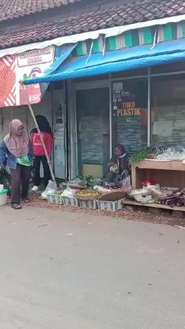 The video shows a small roadside market stand outside a building with a blue awning and the words "SOLUSI BELANJA HARIAN" and "TOKO PLASTIK" printed on it. An older woman wearing a hijab is sitting behind the vegetable stand, arranging her merchandise. There are various vegetables, fruits, and produce items displayed in baskets and on a wooden table. Another woman, wearing a denim jacket and pants, bends down to look at the produce, seemingly interested in buying something. A third woman, dressed in a red sweatshirt and black pants, stands nearby, observing the scene. A motorcycle is parked to the left of the frame. The street appears to be a quiet, possibly rural, area.