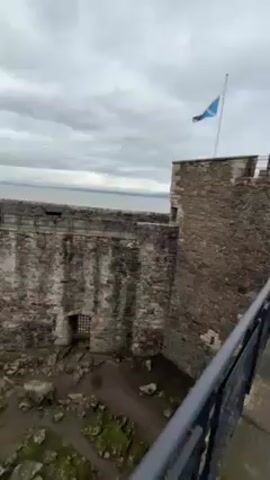 The camera pans from a stone wall with a barred window to a view of a castle in Scotland. The sky is overcast. The Scottish flag is seen on top of a turret in the distance. A stone wall follows the perimeter of the castle grounds. The ground is covered in rocks and what appears to be a layer of green moss. The castle is made of grey stone with small, rectangular windows. A narrow walkway leads towards the building. The narrator mentions that the filming of the show "Outlander" took place at this location.