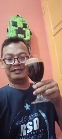 A man wearing glasses and a dark blue t-shirt holds a glass of dark liquid up to the camera. Behind him, a green backpack with a pixelated face design is visible against a peach-colored wall. The man smiles and appears to be speaking.