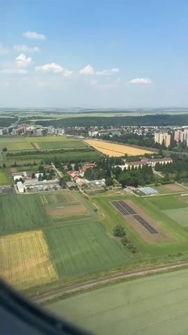 This is an aerial view from an airplane window of agricultural fields and a suburban area. The fields in the foreground are divided into plots of varying colors, suggesting different crops or stages of growth. Some plots are bright green, others are a yellowish-green, and some appear to be bare earth or dry crops. In the middle ground, there are a few buildings, possibly farm structures or research facilities, surrounded by trees. Further in the distance, there is a town or city with residential buildings and some commercial structures. A highway runs parallel to the edge of the town. The sky is blue with scattered white clouds. The airplane's window frame is visible in the foreground, partially obscuring the view. The overall scene depicts a transition from rural agricultural land to a developed urban area.