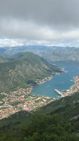 The video captures an aerial view of a scenic bay surrounded by lush green mountains. The initial wide shot shows the vast expanse of the bay and the surrounding mountainous landscape under a partly cloudy sky. A town with numerous buildings, characterized by terracotta roofs, lines the curving shoreline at the base of the mountains.

As the camera slowly zooms in and pans downwards, the focus shifts to the activity within the bay. A large cruise ship, predominantly black with white upper decks and a red and black funnel, is prominently featured, appearing to be docked or anchored close to the town's waterfront. Numerous smaller boats, including speedboats and yachts, are scattered across the turquoise waters of the bay, many leaving white wakes as they move. Some smaller boats are also docked near the shore. The steep slopes of the mountains are covered in dense vegetation, contrasting with the blue water and the urban sprawl of the town. The overall impression is one of a vibrant coastal settlement nestled within a dramatic natural landscape.