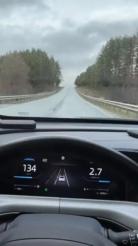 The video shows a driver's perspective from inside a car traveling on a wet, rural road lined with trees. The sky is overcast. The car's digital dashboard displays a speed ranging from 131 to 135 km/h, an engine speed around 2.7-2.8 x1000r/min, and an outside temperature of 4 degrees Celsius. The dashboard also shows a representation of the car and lane assist features. The windshield wipers are active, clearing rain.