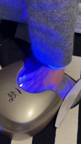 The video shows a person's hand inside a nail polish curing lamp. The lamp is a metallic color and has "Peggy Sage PARIS" printed on it. The hand is illuminated by a bright blue light emanating from the lamp. The person is wearing a gray knitted sweater. The lamp is resting on a black and white striped surface. A nail file is placed next to the lamp. The person's hand is positioned such that the fingernails are directly under the light.