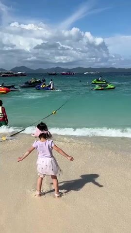 A little girl in a pink hat and a white dress stands on a sandy beach facing the ocean. Her shadow is cast on the sand. She appears to be about to step into the water. In the ocean, several jet skis are visible, as well as a yellow and red inflatable tube. A person in a red life vest is standing in the water near the shore. The water is clear and turquoise, and the sky is blue with white clouds. In the background, there are mountains and other boats.