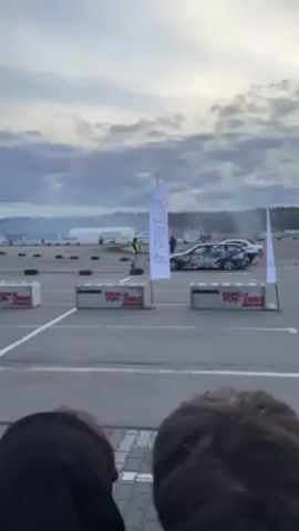 The video shows a drifting event on a paved course marked with white lines, concrete barriers, and stacks of tires. Two cars are performing drift maneuvers, with smoke rising from their tires. One car is dark-colored, the other is white. Several people can be seen watching from behind the barriers, their heads visible in the foreground. A sign on a white pole and another on the concrete barrier advertise "PETER" and "FUN DRIVE". The sky is cloudy and overcast.