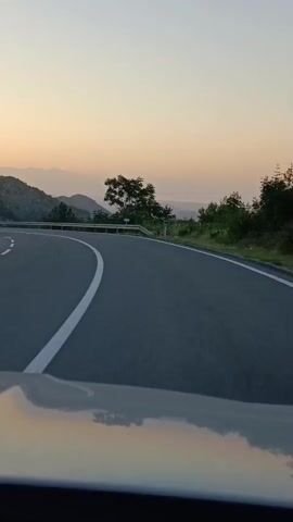 The video shows a first-person perspective from inside a vehicle as it drives along a winding road. The road has a white line marking its edge. On the right side of the road, there is a metal guardrail and trees. Beyond the guardrail, there is a view of rolling hills covered in greenery and trees, with mountains in the distance. The sky is a pale orange and yellow, suggesting either sunrise or sunset. The reflection of the sky and the road is visible on the vehicle's surface in the foreground.