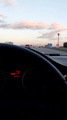 The view from the inside of a moving car shows a highway at dusk. Traffic consists of a variety of vehicles. The sky is partly cloudy, with pinkish hues near the horizon and a pale moon visible.  A road sign in the background is partly visible, featuring Arabic script and the word "GARE." A speed limit sign of 100 is also visible on the right side of the road. The dashboard of the car is in the foreground, with the steering wheel and some illuminated information display visible.  Music is audible.