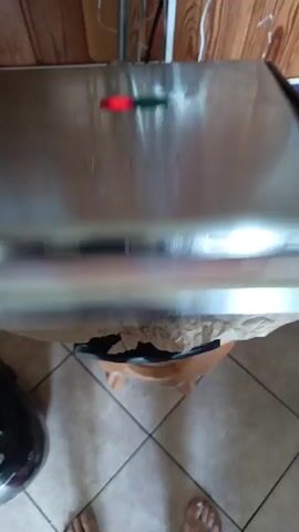 The video shows a close-up, top-down view of a panini press with what appears to be raw meat patties covered in butcher paper. The meat has grill marks on it, indicating it has been pressed or cooked before. The panini press is black and has a ribbed surface. The butcher paper is brown and wrinkled, with some red juices from the meat visible on it. The background is out of focus but appears to be a kitchen or food preparation area, with tiled flooring and wooden cabinets. The lighting is dim.