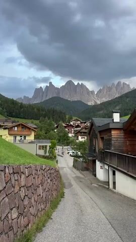 This media file captures a scenic view of a village nestled in the Italian Alps, with the distinctive peaks of the Dolomites forming a dramatic backdrop. The scene is dominated by traditional alpine architecture, featuring wooden houses with pitched roofs and balconies, some adorned with colorful flower boxes. A winding paved road leads through the village, with a few cars visible.

In the foreground, a textured stone wall borders a neatly manicured grassy slope. Trees and dense green forests cover the lower slopes of the mountains, transitioning into the rugged, jagged peaks that pierce a cloudy sky. The weather appears overcast, with dramatic, dark clouds suggesting the possibility of rain or a recent storm, adding a sense of moodiness to the otherwise picturesque landscape.

The time of day is difficult to pinpoint precisely due to the cloudy conditions, but the lighting suggests it is likely late afternoon or early evening. There are no discernible people actively engaged in an event, though a few figures can be seen in the distance walking along the road. The overall impression is one of tranquility and natural beauty, characteristic of a mountain village. The location is identified as St. Magdalena (Santa Maddalena), Italy, a well-known spot for its stunning views of the Geisler/Odle mountain group. No text is visible in the image.