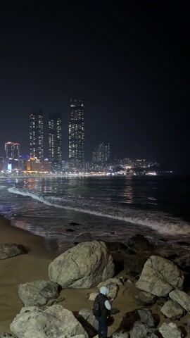 At night, a person on a rocky beach is standing near some rocks, looking at a city across the water. The person is wearing a hoodie, vest, and carrying a backpack. There are large buildings with lit-up windows in the background. The tide is coming in on the beach, with gentle waves washing the shoreline. The overall tone is calm and peaceful.