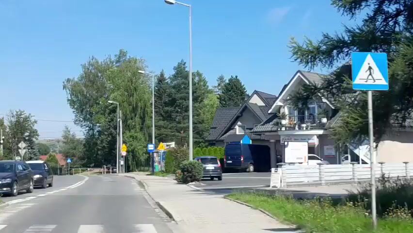 The video shows a street view of a town on a sunny day. There are several buildings, including a motorcycle dealership called CFMOTO PODHALE, which also sells other brands like GOES and TALARIA. The dealership has a large glass facade with various signs advertising motorcycles and accessories. Other businesses visible include MOTObistro and a sign for "Centrum". Several vehicles are present, including cars, motorcycles, and a white van. Some people are seen near the motorcycles, possibly customers or employees. The general environment appears to be a commercial area with trees and a clear blue sky.