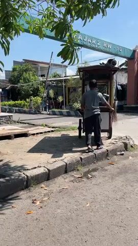 The video shows a man standing behind a food cart. He is wearing a gray t-shirt and black pants. He appears to be tidying up the cart. The food cart has wheels and appears to be made of wood. There are items inside the cart, including what looks like bags of food. There is a broom leaning against the cart. The cart has writing on it that is partially visible. The environment appears to be outdoors, possibly a street or market. There is a sign in the background that says "GADING FAJAR 2". The ground is paved and there is some debris on the ground. The sky is clear and the sun is shining.