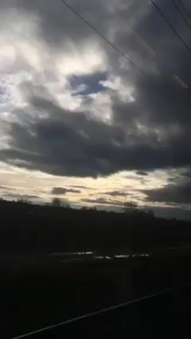 The video depicts a view from a moving train. The camera is pointed out the window. Outside, a field and trees are visible in silhouette against a cloudy sky. The sky is overcast with dark clouds, but there is a break in the clouds allowing some light to shine through. Overhead electrical wires and support poles are visible, passing quickly as the train moves. The overall tone is gloomy due to the dark clouds.