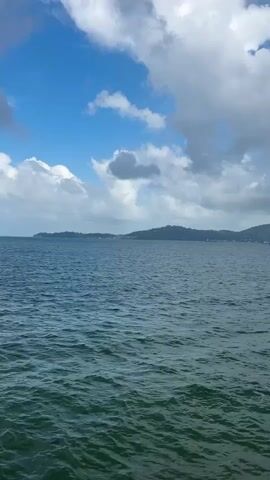A video shows a body of water with a distant coastline under a blue sky with white clouds. The water is choppy with small waves. The coastline is lined with green hills. The sky is bright and partly cloudy.