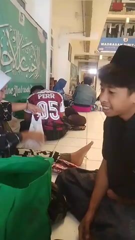 The video shows an indoor hallway with tiled floors and a row of lockers or cubbies on the right side of the frame. A sign above the lockers reads "MADRASAH DINIYAH HATI". People are sitting on the floor, some leaning against a green wall with Arabic calligraphy. In the foreground, a young person wearing a black cap and shirt is looking to the right, smiling. To the left, another person wearing a white hijab and a black and red soccer jersey with "PERR 05" on the back is handling some white plastic bags. Several other people, including a person in a blue headscarf and a person in a grey jacket, are visible sitting on the floor further down the hallway. Green bags and other items are scattered on the floor. The lighting suggests an indoor, possibly institutional setting.