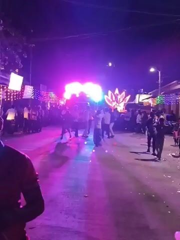 The video shows a street scene at night, illuminated by bright, colorful lights. A crowd of people is gathered, with some individuals dancing and others observing. There are large light displays, including one that resembles a lotus flower, and strings of smaller lights adorning buildings and structures along the street. The overall atmosphere appears festive and energetic. The camera is handheld and moves around, capturing different perspectives of the event. Some people are wearing brightly colored clothing.