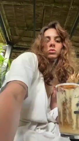 A woman with curly brown hair sits under a thatched roof and stirs a tall glass of iced coffee. The drink has distinct layers of light and dark color, and the glass is branded with a black mountain graphic and the word "Labana". The woman is wearing a loose white shirt.  She uses a black straw to stir the contents of her drink. There are green plants visible in the background.