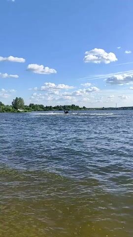 Two jet skis are being ridden on a body of water under a partly cloudy blue sky. The jet skis are moving from left to right across the frame and are kicking up wakes. The shoreline with trees is visible in the background.
